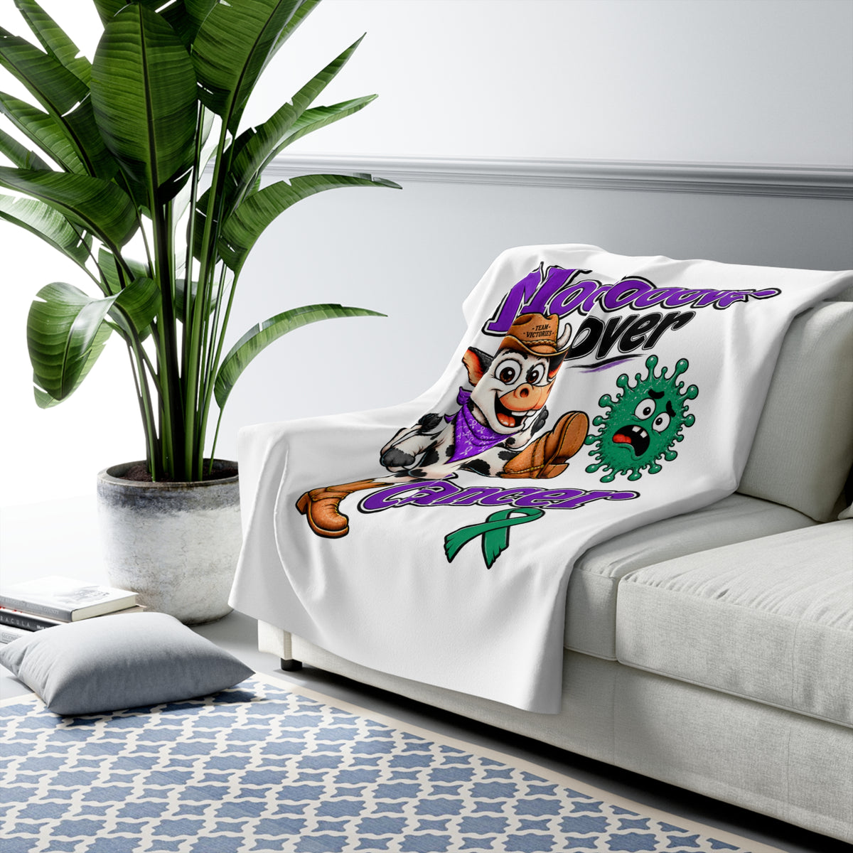 Moooove Over Cancer sherpa blanket draped over a sofa with cow artwork and Liver Cancer awareness ribbon