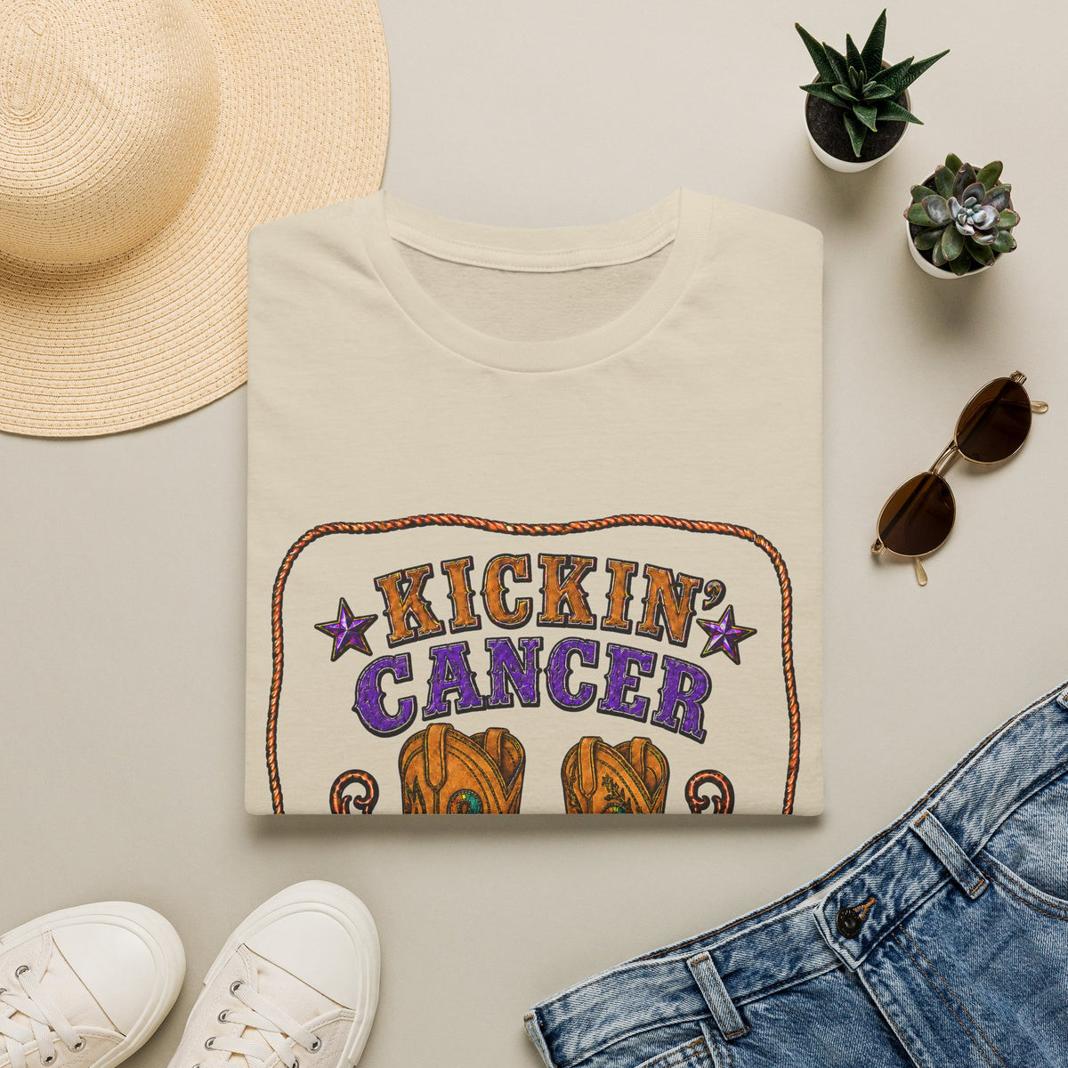 Folded sand-colored Kickin' Cancer T-shirt styled with jeans, sneakers, hat, and sunglasses