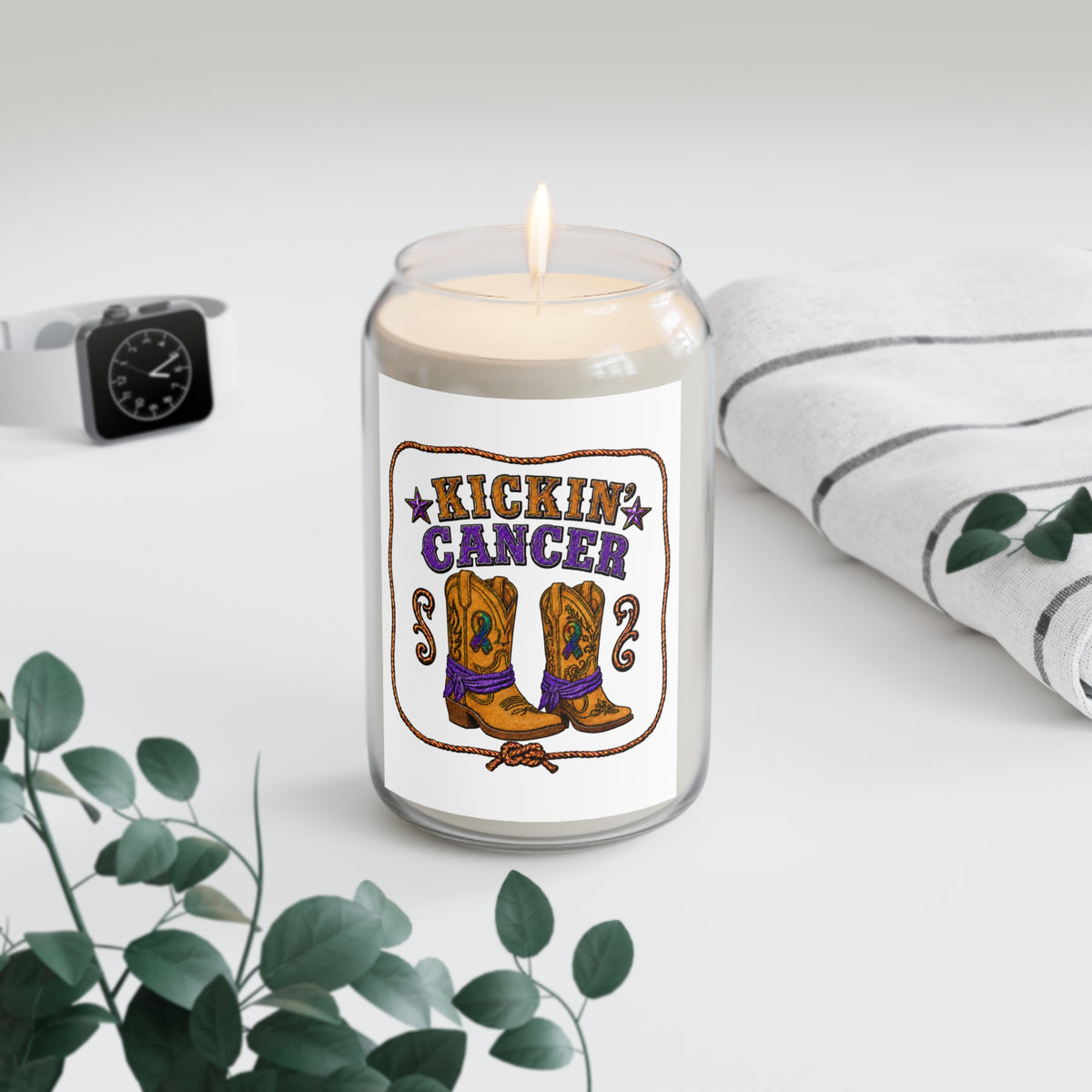 Kickin' Cancer soy candle with western boot artwork beside a clock, houseplant, and folded blanket